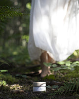 Person in a white dress walking through a forest with a jar of Henua microbiome mask on the ground.