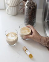 Person holding a glass of coffee on a kitchen counter, Henua eye serum nearby, with a bottle of coffee beans and a jar in the background.