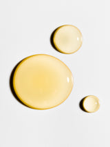Three droplets of yellow liquid Henua oil cleanser on a light gray background